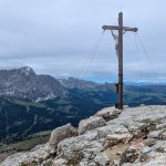 Foto 14: Vrcholový kríž Col de la Pieres na skalnatom temene, v pozadí masív Sassolunga (Langkofel) a údolie Val Gardena.