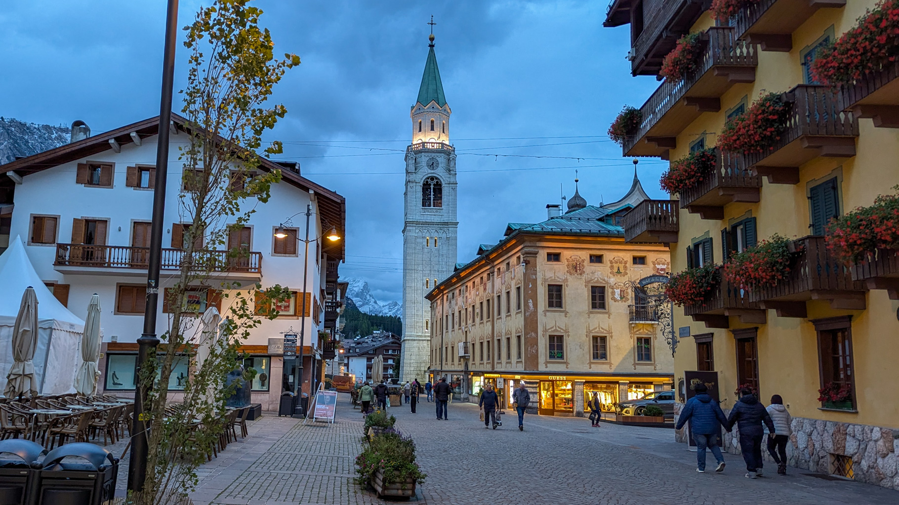 Večerná atmosféra v historickom centre Cortiny d'Ampezzo s dominantnou zvonicou kostola Basilica Minore dei Santi Filippo e Giacomo a elegantnými historickými budovami lemujúcimi Corso Italia.