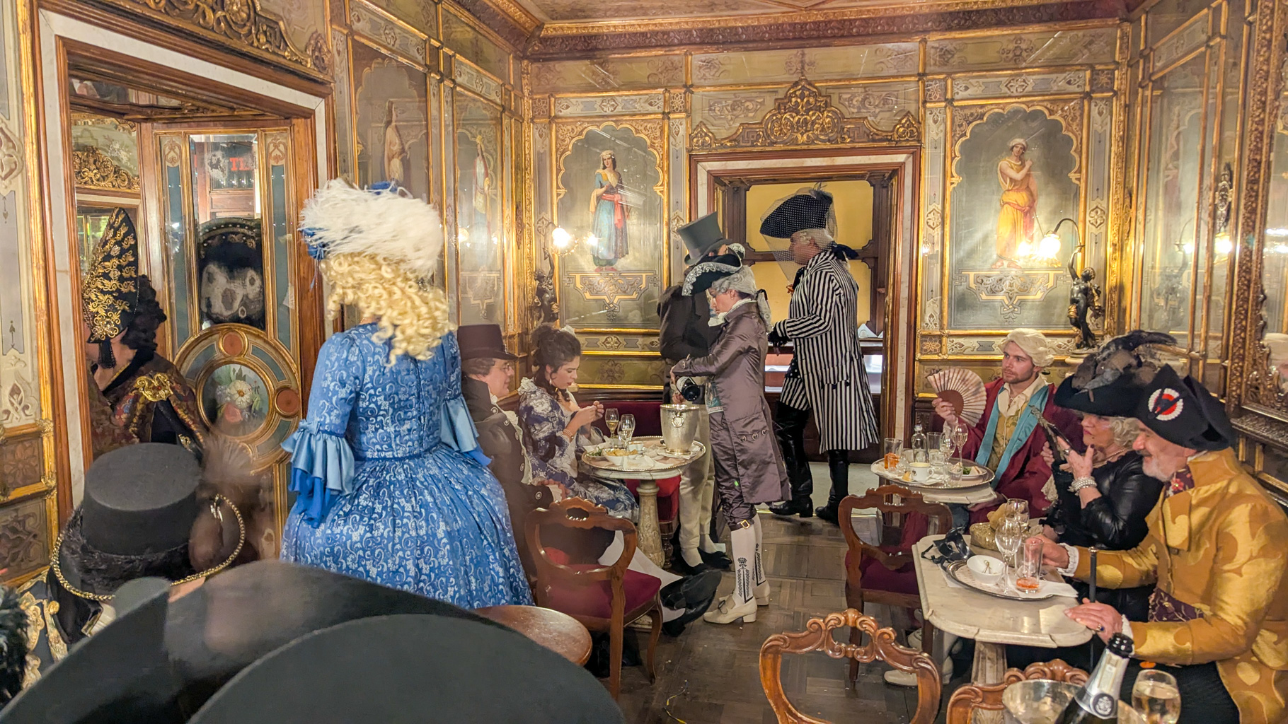 Interiér legendárnej kaviarne Caffè Florian počas benátskeho karnevalu, s návštevníkmi v historických
            kostýmoch. Kaviareň bola založená v roku 1720.
        
