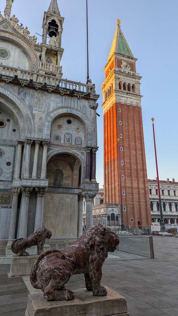 Detail levov a fasády Baziliky svätého Marka so slávnou zvonicou Campanile di San Marco v pozadí za
            ranného svetla.
        