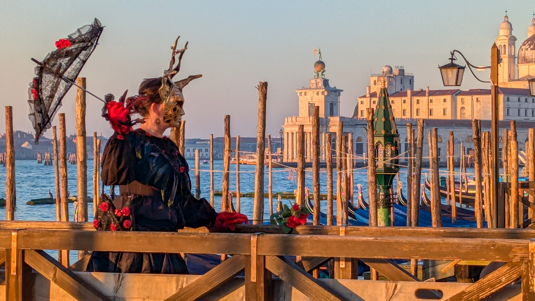Detail karnevalovej postavy s maskou a slnečníkom na móle pri pohľade na Punta della Dogana a Santa Maria
            della Salute.
        