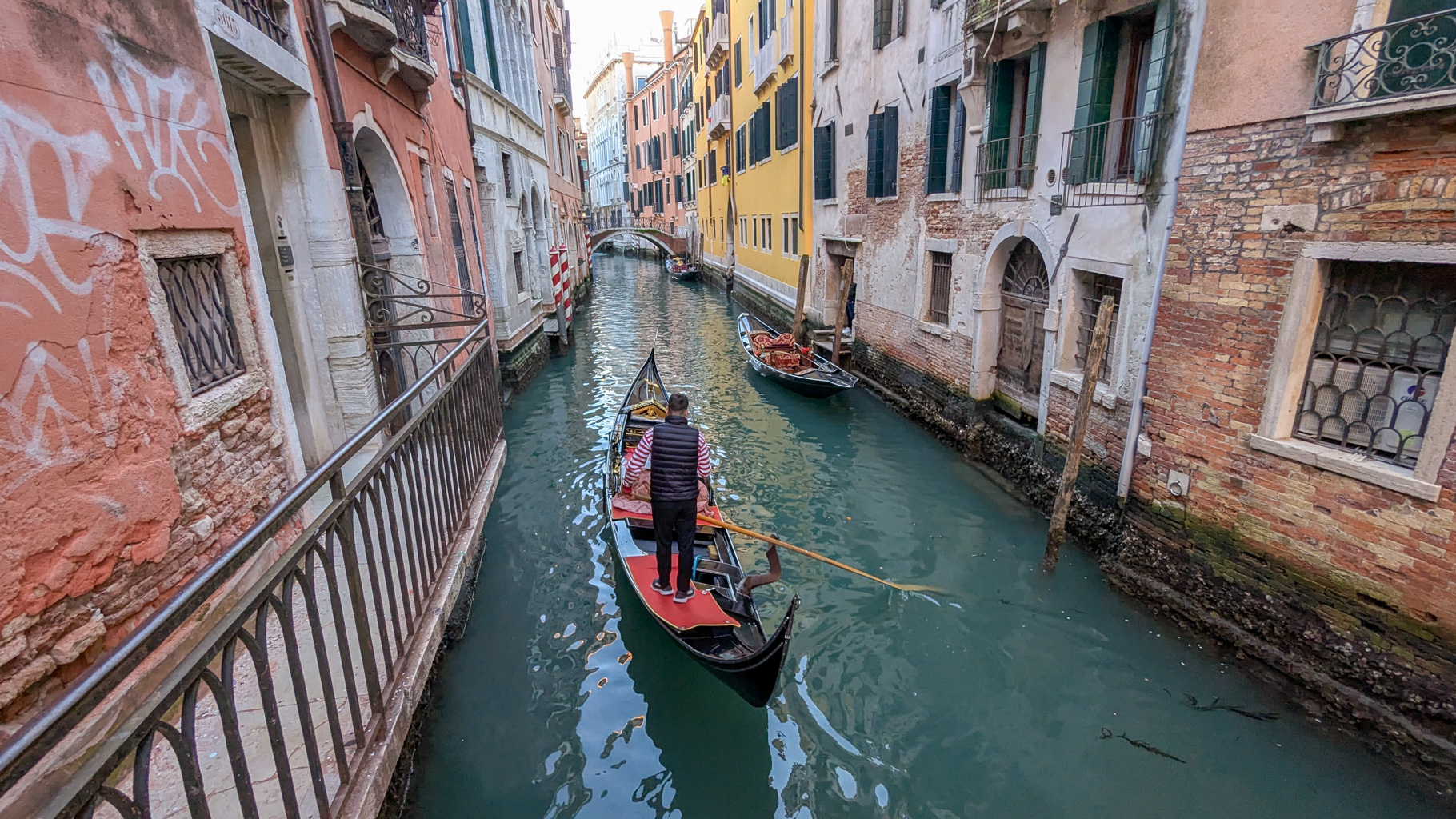 Gondolier vedie tradičnú gondolu cez úzky kanál medzi benátskymi domami. Plavba gondolou je typickou
            atrakciou Benátok.
        